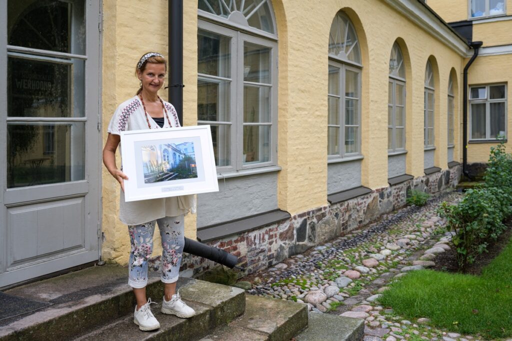 Oven edustalla rappusilla seisova henkilö pitelee kehystettyä kuvaa kädessään, taustalla pihamaa. 